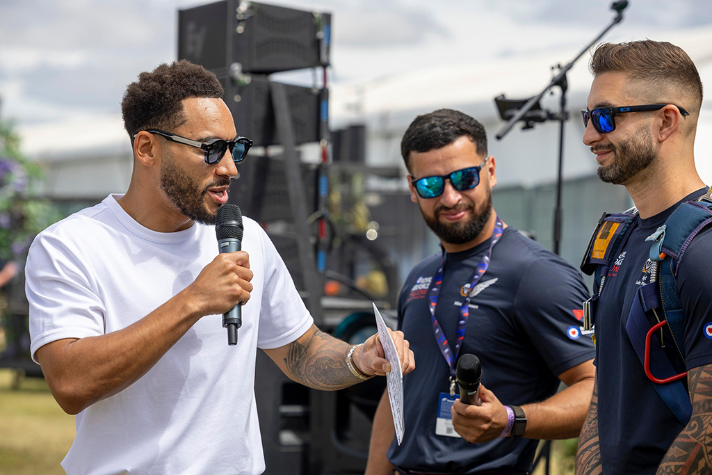 In an outside space, two Royal Air Force Falcons Parachute Display Team members, in conjunction with a compare, stand with microphones answering questions.